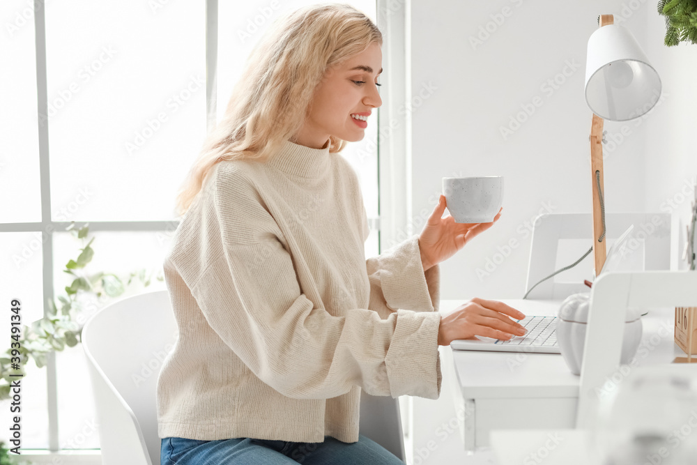 Beautiful young woman in warm clothes drinking tea while using laptop at home Beautiful young woman in warm clothes drinking tea while using laptop at home