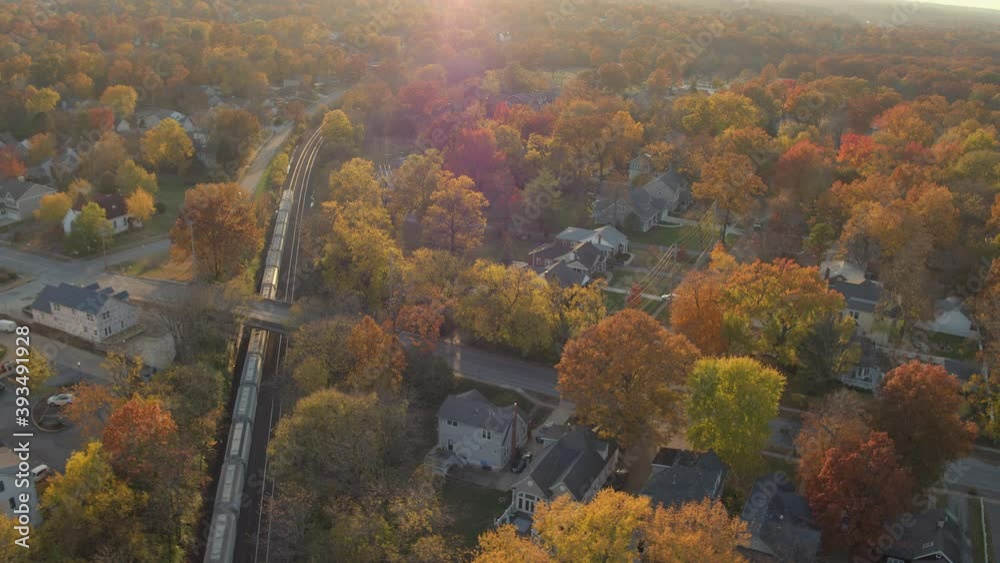 Aerial following a freight train as it passes by a train station in Kirkwood in St. Louis, Missouri on a beautiful Autumn evening at golden hour in November.