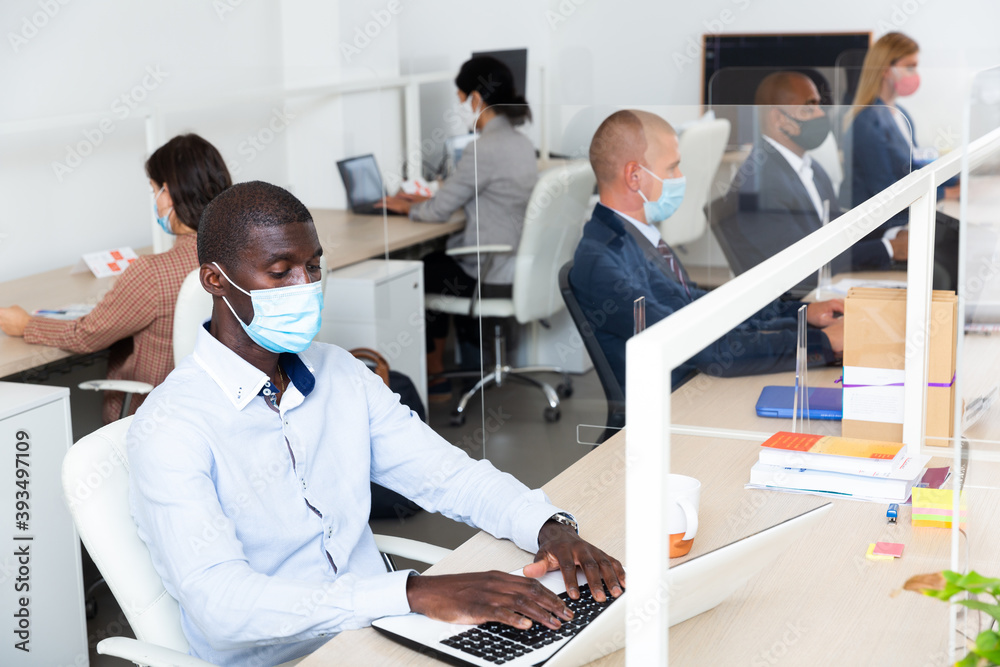 Fototapeta premium Portrait of focused afro american businessman in medical face mask working in open plan office. Pandemic prevention and social distancing concept