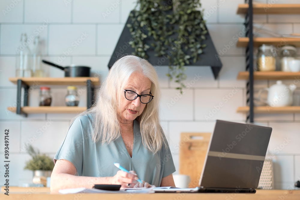 Senior woman using laptop for websurfing in her kitchen. The concept of senior employment, social security. Mature lady sitting at work typing a notebook computer in an home office. Senior woman using laptop for websurfing in her kitchen. The concept of senior employment, social security. Mature lady sitting at work typing a notebook computer in an home office.