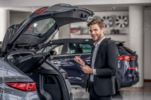 Bearded male standing near open car boot, taking notes