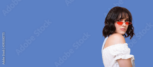 Adorable woman with red glasses is posing in a dress and bare shoulder on a blue studio wall