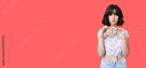 Caucasian woman is doing funny face while holding a tasty donut on a red studio wall with free space