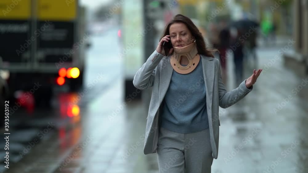 middle-aged businesswoman with cervical collar is walking on street and talking by mobile phone
