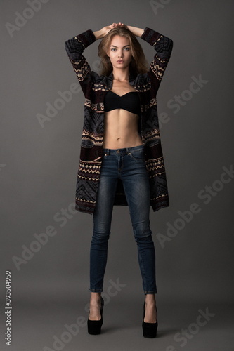 beautiful slender blonde model in a black bra, jeans on the shoulders put on a coat posing on a gray background