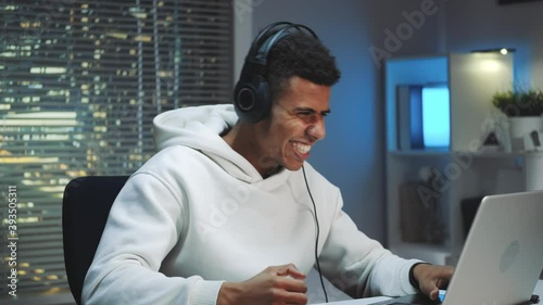 Medium shot of african gamer in headphones winning online game on computer. He is excited and happy. Skyscrapers in the background.