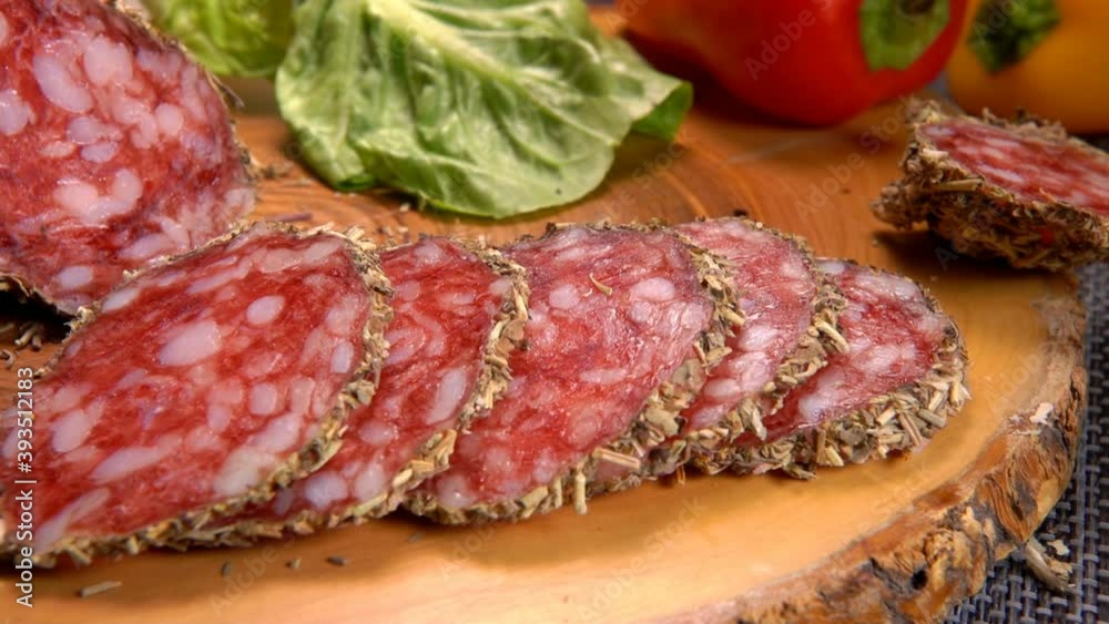 The knife is cutting a delicious dry sausage in herbs on a wooden board on the background of greenery The knife is cutting a delicious dry sausage in herbs on a wooden board on the background of greenery