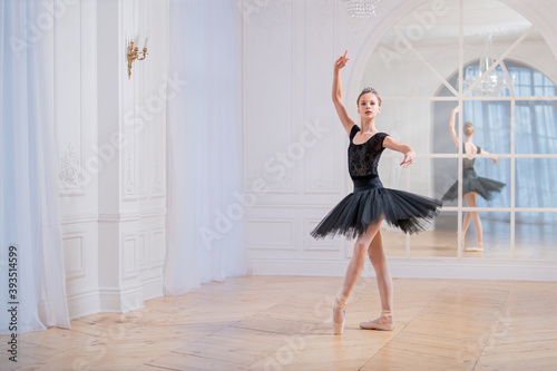 young ballerina in a black tutu is dancing on pointe in large bright hall in front of a mirror. © Maria Moroz