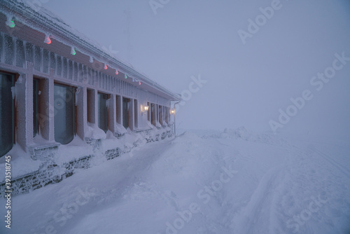 Ski resort in Lapland, Finland. Winter cloudy foggy misty day. Low visibility, silhouettes of people and wooden cabins in the mist. Moody winter cold scene, cafe covered in snow crust 