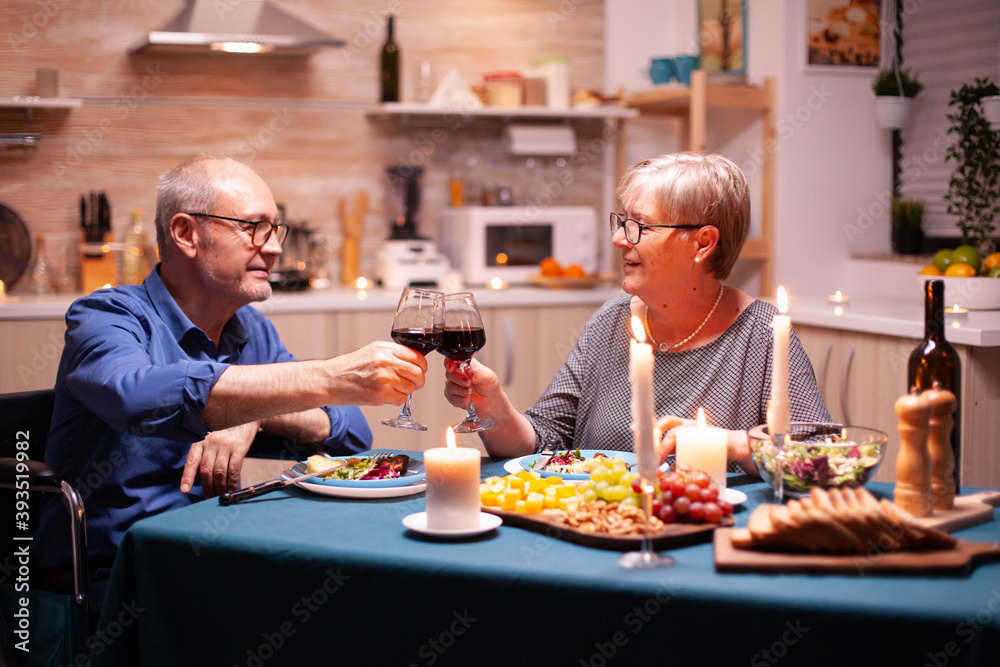 Man in wheelchair dining with wife and toasting using glasses with red wine. Wheelchair immobilized paralyzed handicapped man dining with wife at home, enjoying the meal Man in wheelchair dining with wife and toasting using glasses with red wine. Wheelchair immobilized paralyzed handicapped man dining with wife at home, enjoying the meal