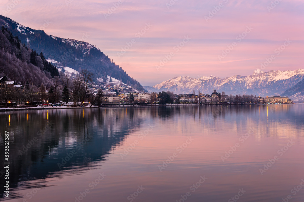 Zell am See in winter evening. View of Lake Zell, town, mountains and snow with reflections in water. Alpine town at purple dusk. Famous ski resort in Alps, Austria. Winter wonderland for travel