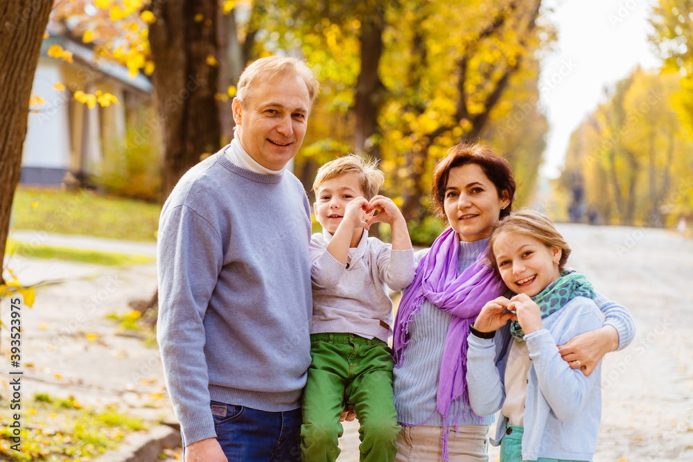 Fototapeta premium A Family of four, mom, dad, son and daughter showing heart with hands enjoying sunny weather golden leaves in autumn park