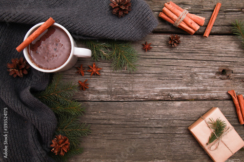 Fototapeta Naklejka Na Ścianę i Meble -  Mug of hot chocolate, spices, grey knitted scarf, fir tree branches, cones, craft gift on rustic wooden table background. Winter and Christmas concept. Spicy cocoa, New Year, top view, copy space