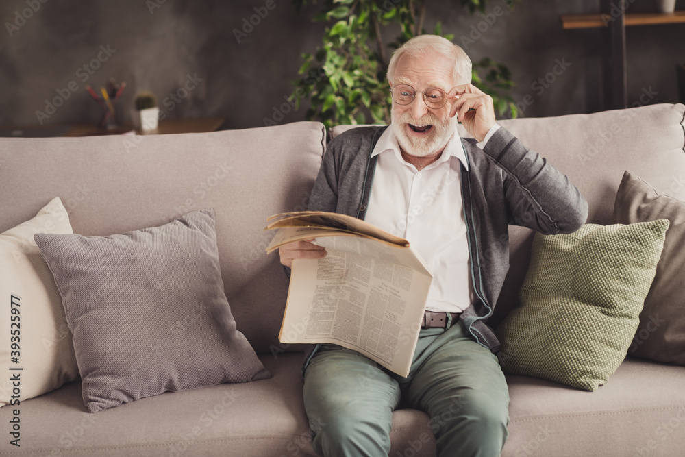 Photo of smiling pensioner wear grey cardigan sitting couch reading newspaper arm glasses indoors house flat