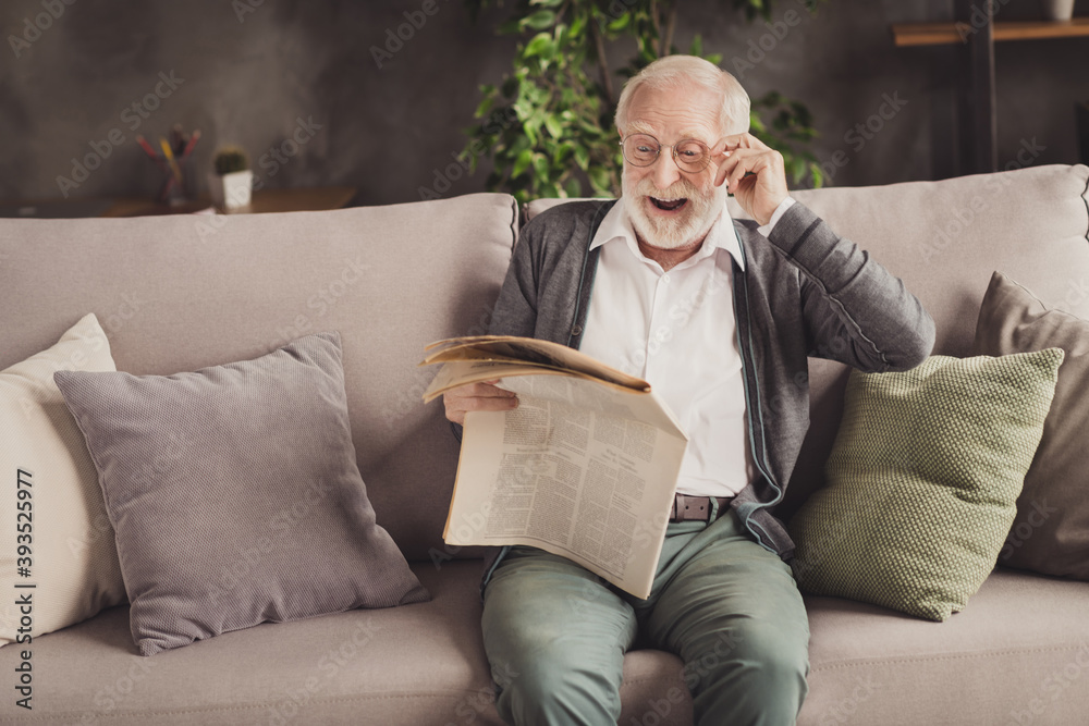 Fototapeta premium Photo of smiling pensioner wear grey cardigan sitting couch reading newspaper arm glasses indoors house flat