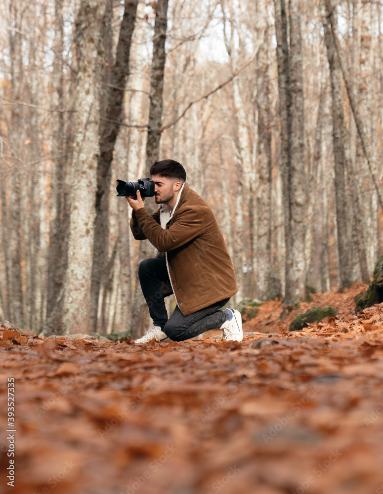 Caucasian boy in beautiful forest with red leaves taking a picture with professional reflex camera photography student with brown jacket, white t-shirt and red scarf, it's cold Caucasian boy in beautiful forest with red leaves taking a picture with professional reflex camera photography student with brown jacket, white t-shirt and red scarf, it's cold