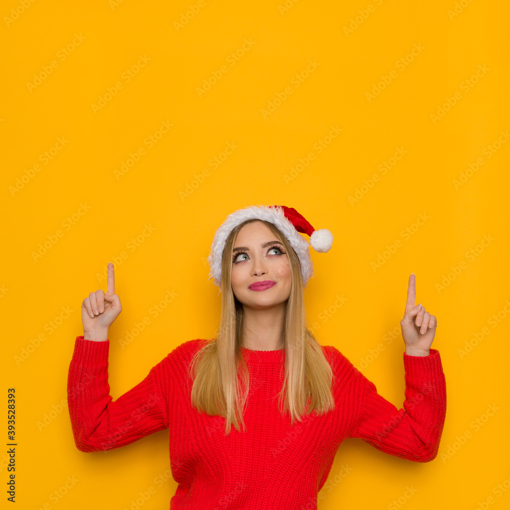 Cute Blond Woman In Santa Hat Is Pointing Up And Looking. Cute Blond Woman In Santa Hat Is Pointing Up And Looking.