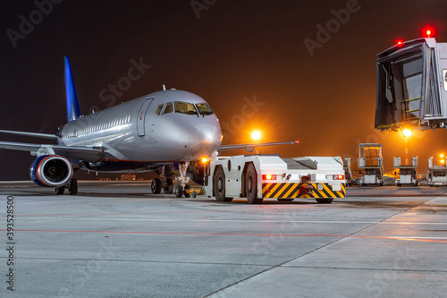 Tow tractor pushes passenger aircraft from boarding bridge at night airport apron