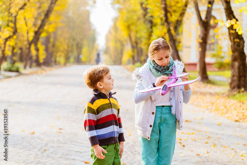 Little girl and boy,launches toy plane, into the air against background of autumn park. Children playing toy plane. Cute sister and brother, stands on track perspective, launches purple toy plane.