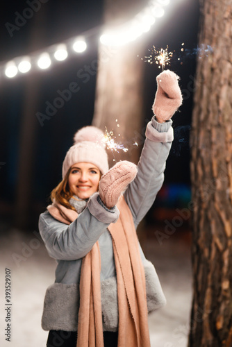 Christmas sparkles in hands. Cheerful young woman celebrating holding sparkles in the winter forest. Festive garland lights. Christmas, new year. 