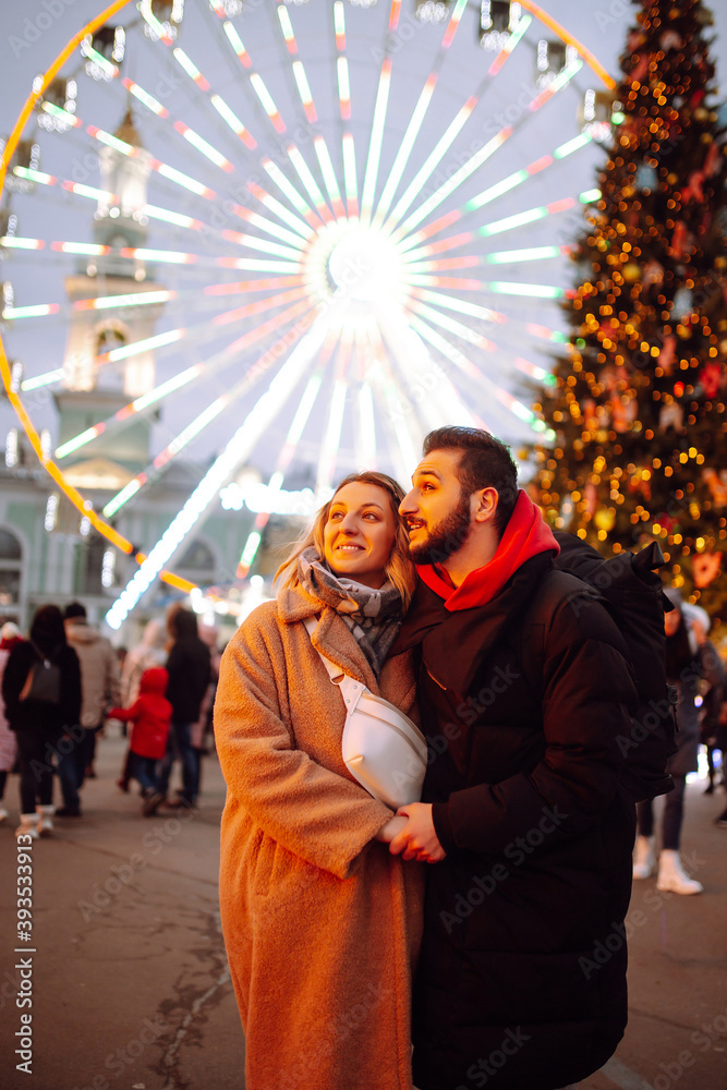 Happy couple in love walking on Christmas market at evening. Young woman and man at festive street market  enjoying winter moments. Lights around. Christmas, New Year.