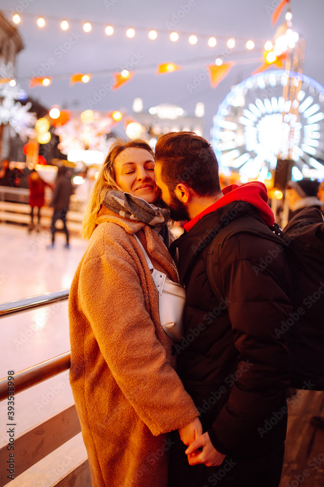 Happy couple hugging and kissing  near skating rink on Christmas market. Young woman and man at festive street market enjoying winter moments. Lights around. Christmas, New Year.