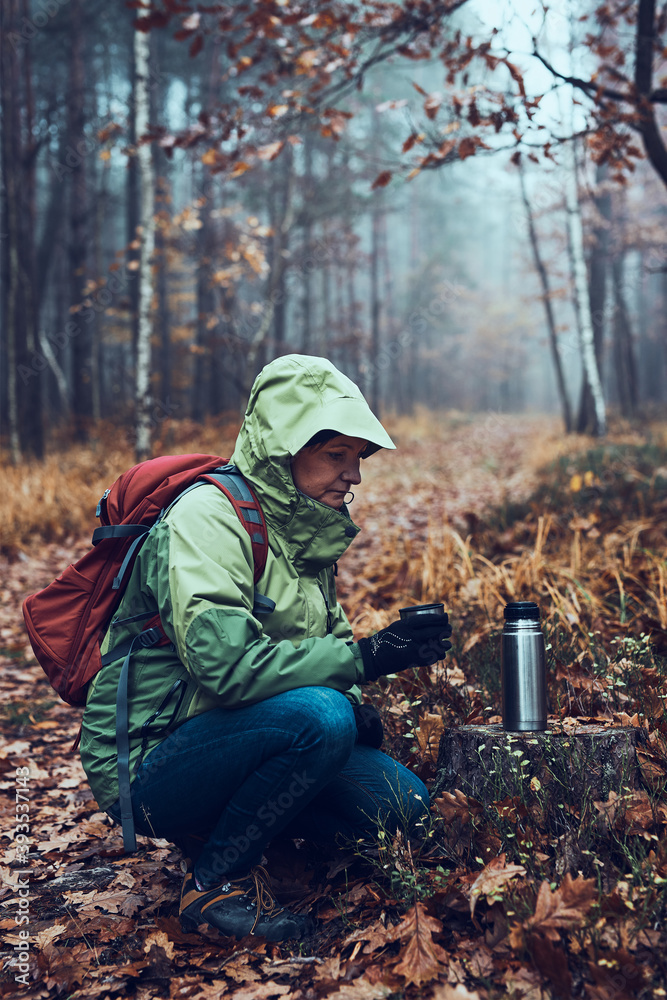 Woman with backpack having break during autumn trip drinking a hot drink from thermos flask on autumn cold day. Active middle age woman wandering around a forest actively spending time Woman with backpack having break during autumn trip drinking a hot drink from thermos flask on autumn cold day. Active middle age woman wandering around a forest actively spending time