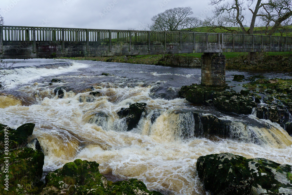 Rocky white river water rapids with a wooden footbridge over it, Linton Falls, Grassington, Yorkshire, UK. Rocky white river water rapids with a wooden footbridge over it, Linton Falls, Grassington, Yorkshire, UK.