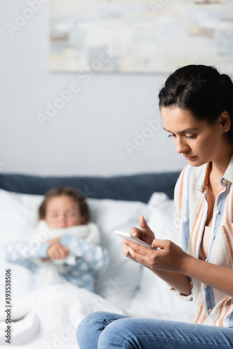mother messaging on mobile phone near diseased daughter lying in bed on blurred background