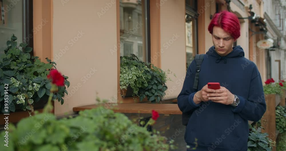 Guy in dark hoodie with bag on shoulder using mobile phone while standing at street. Male millennial hipster with dyed red hair browsing internet while typing and looking at phone screen. Guy in dark hoodie with bag on shoulder using mobile phone while standing at street. Male millennial hipster with dyed red hair browsing internet while typing and looking at phone screen.