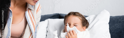 woman sitting near diseased daughter sneezing in paper napkin while lying in bed, banner