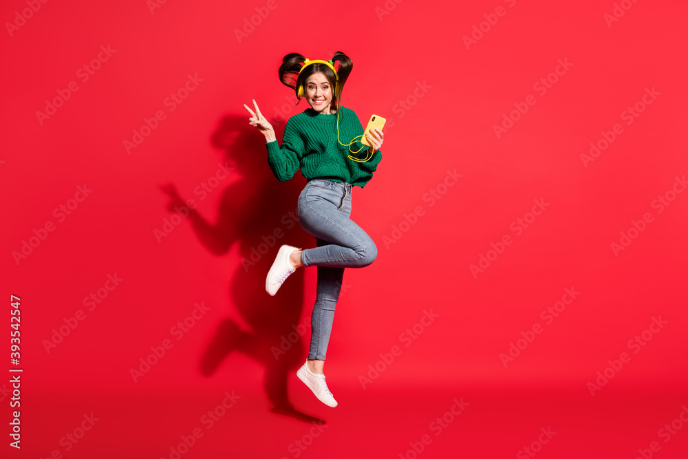 Full size photo of young cheerful positive girl show v-sign listen music in headphones hold mobile isolated on red color background