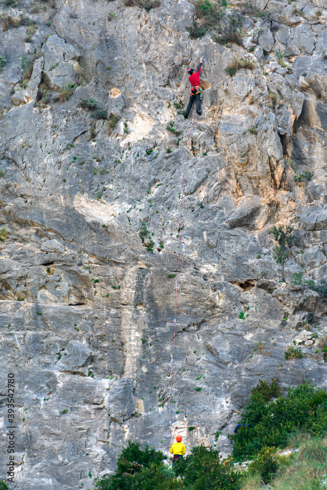 Two climbers, one climbing and the other securing, on a mountain with a vertical wall practicing sports.