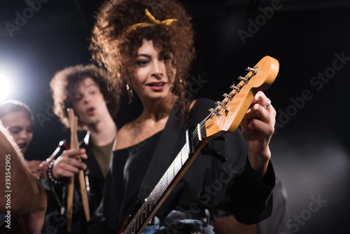 Curly woman touching and looking at electric guitar, while standing near musician pointing with finger on blurred background