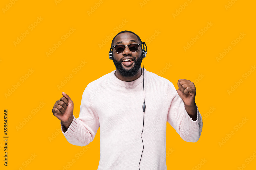Cheerful African American attractive man in eyeglasses enjoys music podcast in headphones, listens audio in music app and dance against orange isolated background in a fanny mood