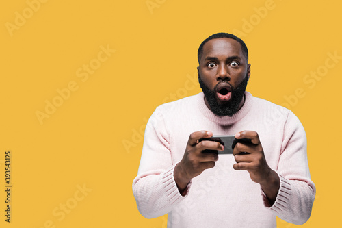 Excited african american man in casual wear is standing on isolated orange background holds his mobile phone in hands and looking surprised at the camera