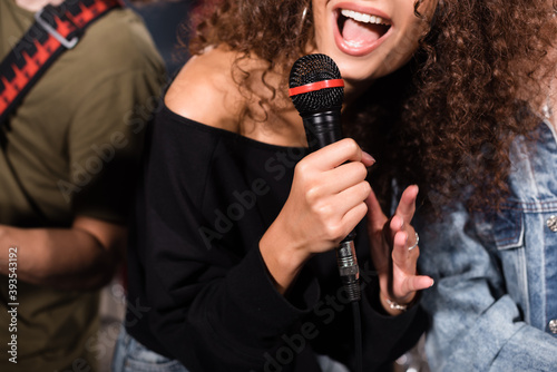 Cropped view of curly female vocalist with microphone singing near rock band musicians on blurred background