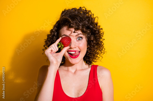 Closeup portrait photo of charming tricky young girl hand hold strawberry cover eye stick out tongue lick lips wear red singlet unclothed shoulders isolated bright yellow color background