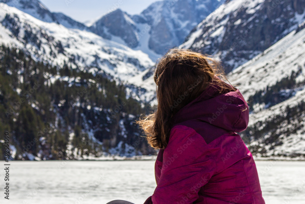 Beautiful young woman sitting next to stunning winter landscape. Concept of travel and adventure at the frozen Gaube Lake (Lac de Gaube) in the French Pyrenees, Cauterets, France.