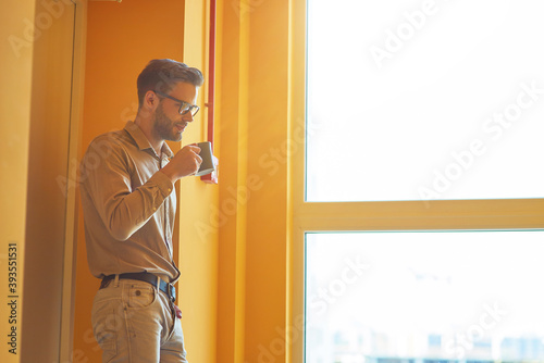 Resting at work. Young handsome businessman drinking coffee or tea while standing in the office and looking out the window