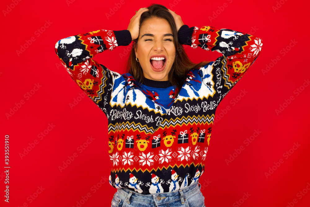 Shocked panic Young beautiful Caucasian woman wearing Christmas sweater against red wall,  holding hands on head and screaming in despair and frustration.