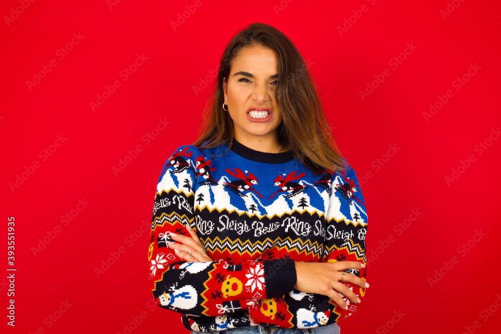Mad crazy Young beautiful Caucasian woman wearing Christmas sweater against red wall,  clenches teeth angrily, being annoyed with coming noise. Negative feeling concept.