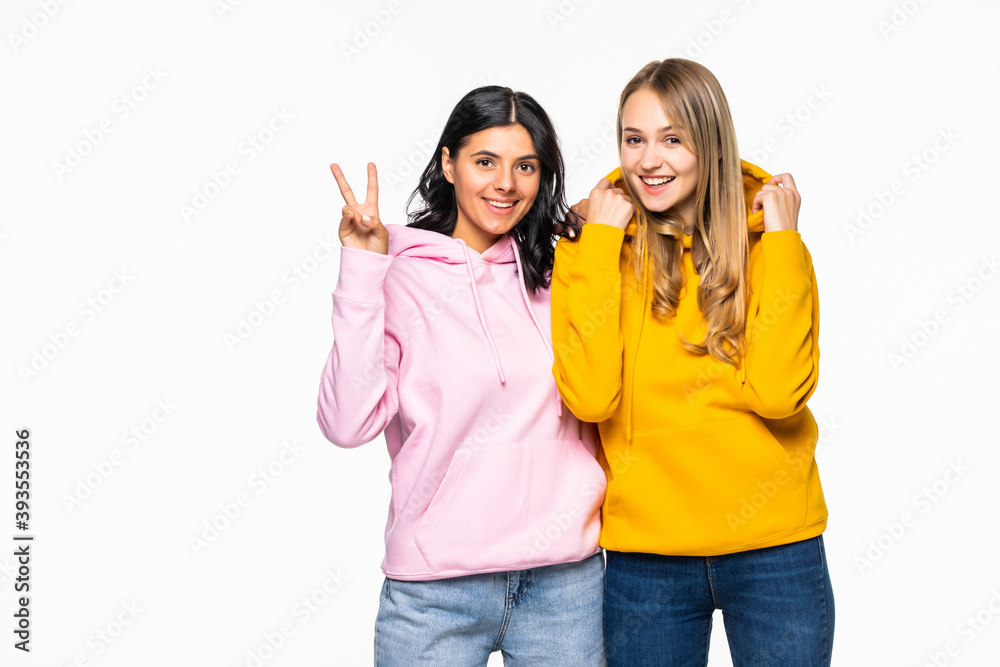 Pretty two girlfriends women showing v-sign symbols wearing casual bright hoodies and jeans isolated white background Pretty two girlfriends women showing v-sign symbols wearing casual bright hoodies and jeans isolated white background