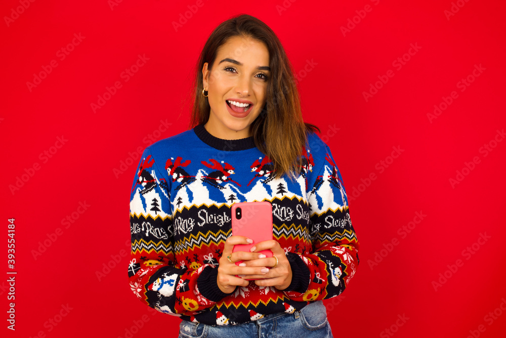 Smiling young beautiful Caucasian woman wearing Christmas sweater against red wall, friendly and happily holding mobile phone taking selfie in mirror. Smiling young beautiful Caucasian woman wearing Christmas sweater against red wall, friendly and happily holding mobile phone taking selfie in mirror.
