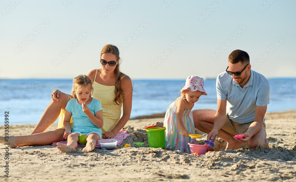 Fototapeta premium family and leisure concept - happy father, mother and two little daughters playing with sand toys on summer beach