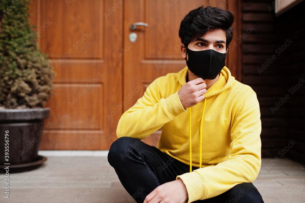 Urban young hipster indian man in a fashionable yellow sweatshirt. Cool south asian guy wear hoodie and black face protect mask, during new normal. Urban young hipster indian man in a fashionable yellow sweatshirt. Cool south asian guy wear hoodie and black face protect mask, during new normal.