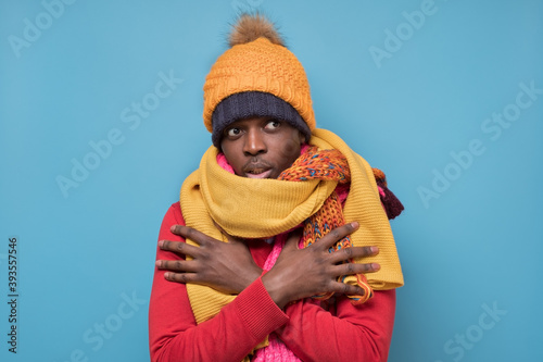 Funny african american mature man in yellow scarf and hat freezing in warm clothing. Studio shoot on blue wall. photo