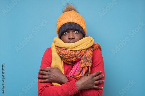 Funny african american mature man in yellow scarf and hat freezing in warm clothing. Studio shoot on blue wall. photo