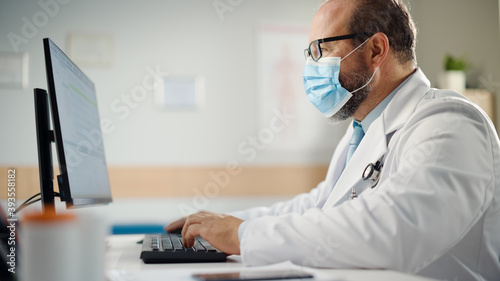 Hispanic Doctor's Office: Experienced Physician Wearing Face Mask Sitting at His Desk Working on Personal Computer. Medical Health Care Specialist Filling Prescription Forms, Checking Test Results