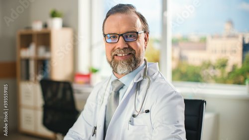 In a Doctor's Consultation Room: Experienced Happy Physician Wearing Glasses, Sitting at His Desk, Smiling at the Camera. Health Care Specialist Checking Test Results. Portrait Shot.